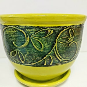 Può includere: Un vaso di ceramica giallo con un motivo floreale verde. Il vaso ha un sottovaso abbinato.