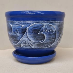 Może przedstawiać: Biały ceramiczny kubek z czarnym uchwytem i napisem "But First, Coffee" w stylu odręcznym. Kubek stoi na białej powierzchni. Kubek ma 10 cm wysokości.