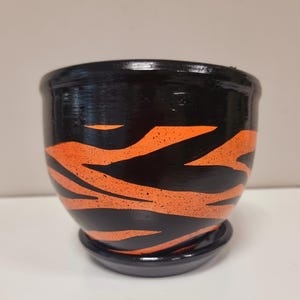 以下が含まれることがあります： 黒いセラミック植木鉢と、お揃いの受け皿。 植木鉢は、大胆なオレンジ色のトラ縞模様が特徴です。 鉢は丸く、縁がわずかにフレアしており、屋内または屋外の植物に適しています。