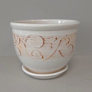 May include: A white ceramic planter with a matching saucer. The planter features a decorative band of brown, stylized floral designs. The pot has a rounded shape and a wide rim. The design is suitable for indoor or outdoor use.