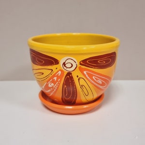 Pode incluir: Um vaso de flores de cerâmica pintado à mão com um pires a condizer. O vaso é amarelo com um design floral estilizado em tons de laranja, castanho e branco. O vaso tem aproximadamente 10 cm de diâmetro e 7,6 cm de altura.