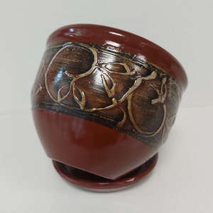 Könnte beinhalten: Ein Terrakotta-Blumentopf mit braunem und goldenem Blumendesign. Der Topf hat ein Abzugsloch und eine passende Untertasse.