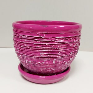 Può includere: Un vaso di fiori in ceramica magenta con un sottovaso abbinato. Il vaso presenta un design a righe orizzontali strutturato con accenti bianchi. Il vaso rotondo è adatto per uso interno o esterno.