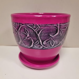 Puede incluir: Una maceta de cerámica rosa con un diseño floral decorativo. La maceta tiene un platillo a juego.