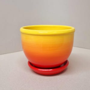 May include: A yellow, orange, and red ombre ceramic planter with a matching saucer. The planter is round and has a smooth, glossy finish.
