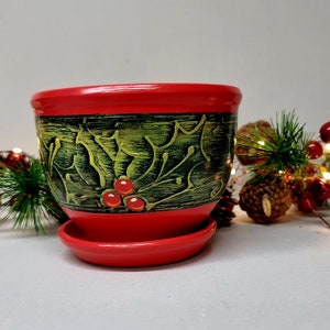 以下が含まれることがあります： 赤い緑のセラミックプランターに、ヒイラギの葉のデザインと赤いベリーが描かれています。プランターには受け皿が付いています。