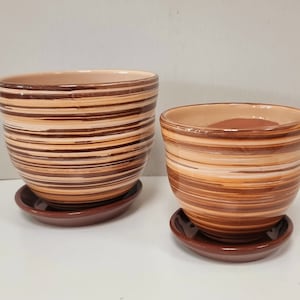 Könnte beinhalten: Zwei Keramik-Blumentöpfe mit passenden Untertassen. Die Töpfe sind braun und beige mit horizontalen Streifenmustern. Die Untertassen sind einfarbig dunkelbraun. Die Töpfe sind unterschiedlich groß.