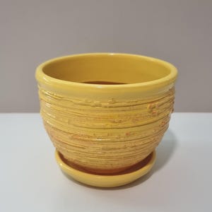 Könnte beinhalten: Gelber Keramik-Blumentopf mit passender Untertasse. Der Topf hat ein strukturiertes, horizontal gestreiftes Design mit einer leicht rauen Oberfläche. Die Farbe ist ein helles, sonniges Gelb. Der Topf hat einen Durchmesser von ca. 13 cm.