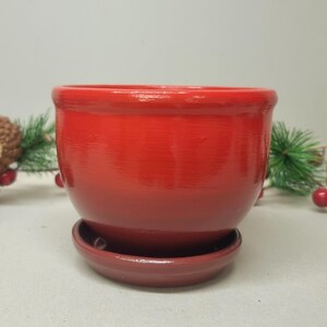 May include: A red ceramic planter with a matching saucer. The planter has a rounded shape and a smooth, glossy finish.