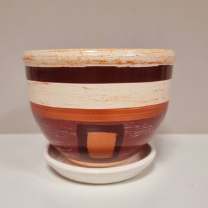 May include: A white ceramic planter with a brown, orange, and white striped design. The planter has a matching saucer.