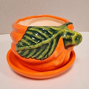 May include: Orange ceramic pumpkin-shaped plant pot with a matching saucer. The pot features a green leaf detail and a cream-coloured interior. The design is reminiscent of autumn and harvest themes.