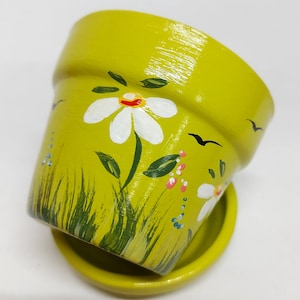 May include: A small, green ceramic planter with a painted design of white daisies, green leaves, and pink and blue flowers. The planter has a matching saucer.