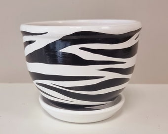 May include: A white ceramic planter with a black and white zebra print design. The planter has a matching saucer.