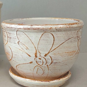 Puede incluir: Una maceta de cerámica blanca con un platillo a juego. La maceta presenta un acabado envejecido y un diseño floral marrón. La maceta es redonda con una amplia abertura, adecuada para plantar flores o hierbas.