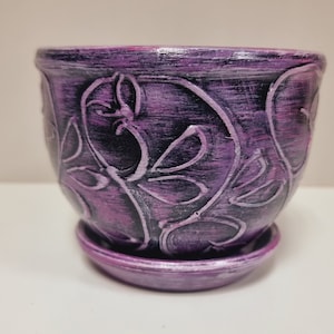 May include: Purple ceramic planter with a decorative floral design. The planter has a matching saucer.