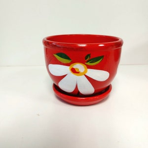 Może przedstawiać: Czerwona ceramiczna doniczka z pasującym spodkiem. Doniczka jest pomalowana w duży biały stokrotka z żółtym środkiem i zielonymi liśćmi. Przeznaczona do użytku wewnątrz i na zewnątrz.