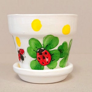 Puede incluir: Una maceta de cerámica blanca con su plato a juego, decorada con tréboles de cuatro hojas verdes pintados a mano, mariquitas rojas con puntos negros y lunares amarillos alrededor del borde superior. El diseño está en el exterior.