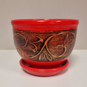 Könnte beinhalten: Ein roter Keramiktopf mit braunem Blumendesign und einem passenden Untersetzer. Der Topf ist perfekt für kleine Pflanzen oder Sukkulenten.