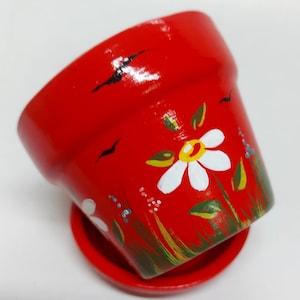 Peut inclure: Pot de fleurs en terre cuite rouge avec une marguerite blanche et des feuilles vertes peintes sur le côté. Le pot a une soucoupe assortie.