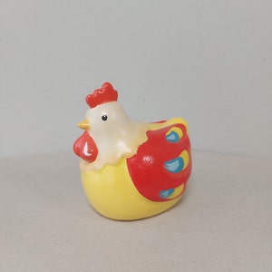 Op de afbeelding: Een keramiek kippenbeeldje met een geel lichaam, rode vleugels en blauwe accenten. De kip heeft een wit hoofd met een rode kam en een zwart oog.