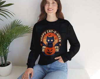 Könnte beinhalten: Ein schwarzes Langarm-Sweatshirt mit einem Grafikdruck einer schwarzen Katze, die auf einem Kürbis sitzt, flankiert von zwei Totenköpfen und Herbstblättern, mit dem Text "HAPPY CAT-O-WEEN" in Orange und Weiß darüber.
