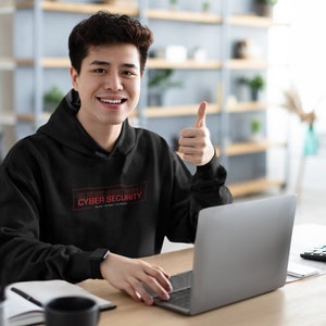 Op de afbeelding: Een jonge persoon draagt een zwarte hoodie met de tekst "ALL PROGRAMS ARE SUSPECT" en "CYBER SECURITY" in rood. Ze zitten aan een bureau met een laptop en geven een duim omhoog.