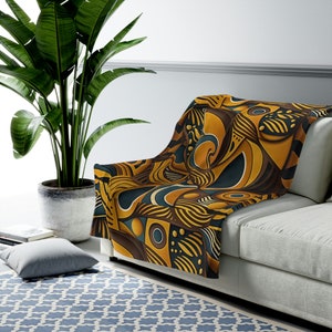 Può includere: Una coperta con un motivo geometrico in tonalità di marrone, giallo e blu. La coperta è drappeggiata su un divano.