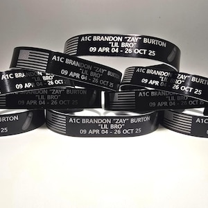 May include: A collection of black silicone wristbands featuring a white American flag graphic and the printed text A1C BRANDON ZAY BURTON LIL BRO 09 APR 04 - 26 OCT 25. These memorial bracelets are stacked together in a group, displaying the same text.