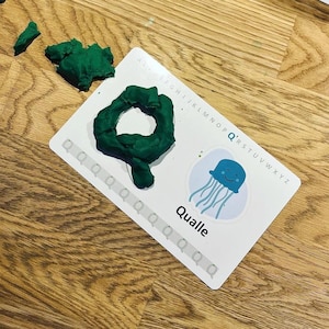K&ouml;nnte beinhalten: Eine wei&szlig;e Lernkarte mit dem Buchstaben Q, einer blauen Quallen-Illustration und dem Wort Qualle. Eine dunkelgr&uuml;ne Knetmasse in Form des Buchstabens Q liegt auf der Karte, daneben befinden sich weitere Knetst&uuml;cke.