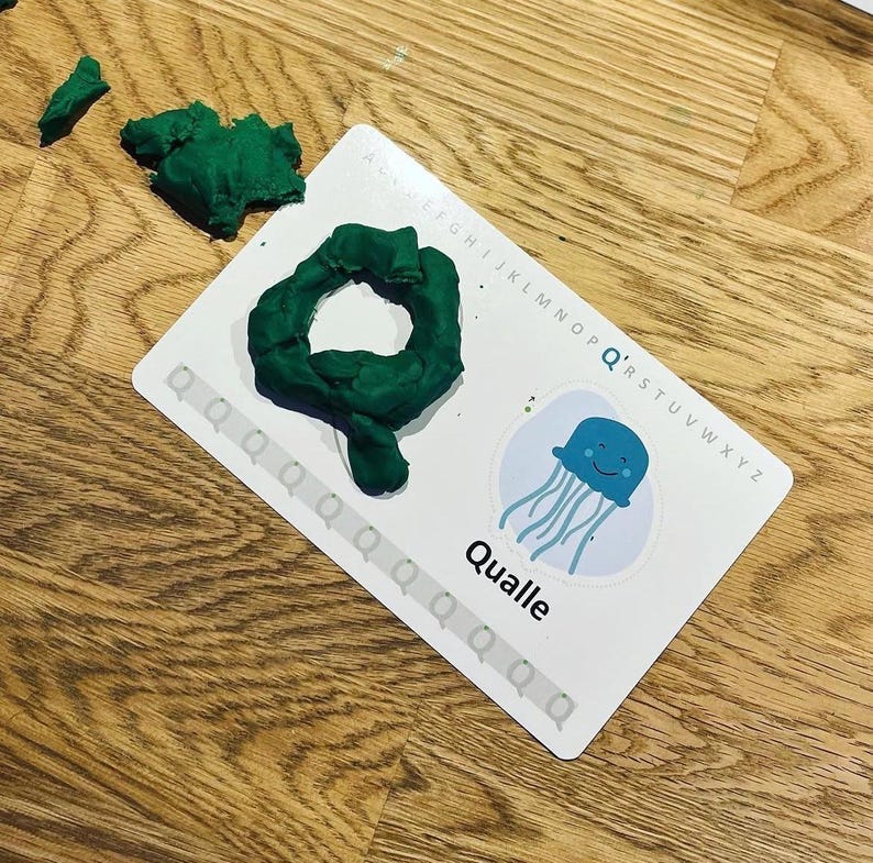 K&ouml;nnte beinhalten: Eine wei&szlig;e Lernkarte mit dem Buchstaben Q, einer blauen Quallen-Illustration und dem Wort Qualle. Eine dunkelgr&uuml;ne Knetmasse in Form des Buchstabens Q liegt auf der Karte, daneben befinden sich weitere Knetst&uuml;cke.
