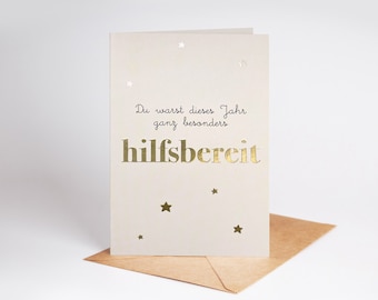 May include: A cream-colored greeting card with gold foil text and star accents. The card reads "Du warst dieses Jahr ganz besonders hilfsbereit" in elegant script and bold capital letters. The card is paired with a brown envelope.