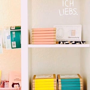 K&ouml;nnte beinhalten: Ein wei&szlig;es Regal mit Stapeln rechteckiger Gegenst&auml;nde. Im oberen Fach befindet sich ein Stapel pfirsichfarbener Artikel und eine Karte mit dem Text "ICH LIEBS." und "&Uuml;berraschung". Im unteren Fach stehen Stapel mit gelben und t&uuml;rkisen Artikeln.