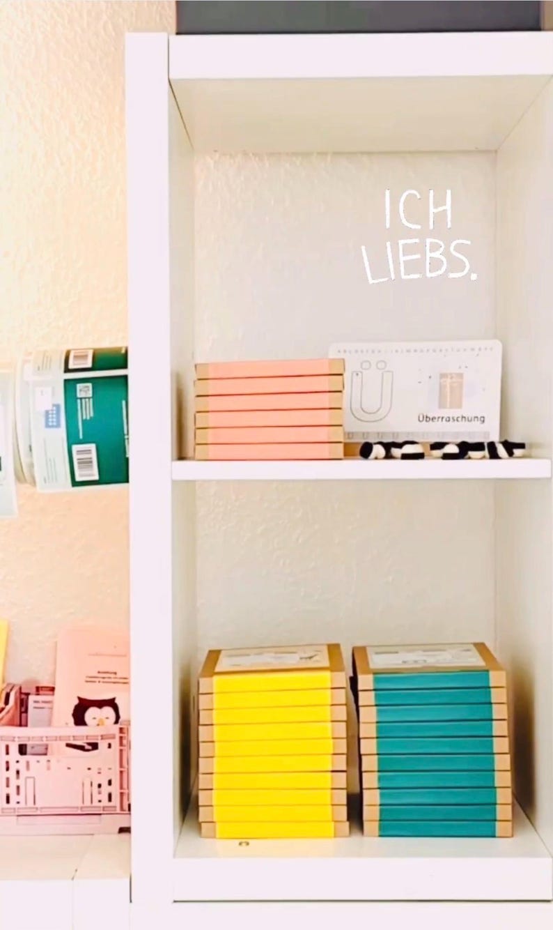 K&ouml;nnte beinhalten: Ein wei&szlig;es Regal mit Stapeln rechteckiger Gegenst&auml;nde. Im oberen Fach befindet sich ein Stapel pfirsichfarbener Artikel und eine Karte mit dem Text "ICH LIEBS." und "&Uuml;berraschung". Im unteren Fach stehen Stapel mit gelben und t&uuml;rkisen Artikeln.