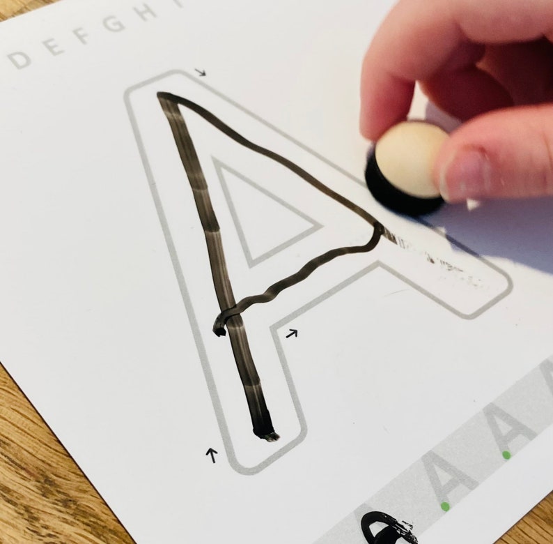 K&ouml;nnte beinhalten: Ein wei&szlig;es Lernarbeitsblatt mit einem gro&szlig;en umrandeten Buchstaben A und Richtungspfeilen zum Nachspuren. Der Buchstabe ist teilweise mit schwarzer Tinte ausgef&uuml;llt. Ein kleines, rundes, helles Holzwerkzeug mit schwarzer Basis liegt auf dem Papier.