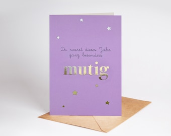 May include: A lavender greeting card with gold foil lettering and star accents. The card reads "Du warst dieses Jahr ganz besonders mutig" in gold foil. The card is paired with a brown envelope.