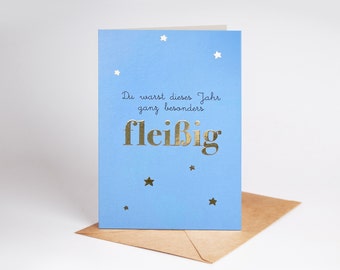 May include: A light blue greeting card with gold foil stars and the German text "Du warst dieses Jahr ganz besonders fleißig" printed in a mix of black script and large gold foil lettering, accompanied by a brown paper envelope.