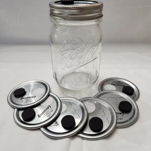 Könnte beinhalten: Ein Satz von sechs silbernen Einmachdeckeln mit schwarzen Gummidichtungen. Die Deckel haben die Aufschrift "Canning" und "Mason". Die Deckel sind in einem Kreis um ein klares Glasgefäß mit einem silbernen Deckel angeordnet.