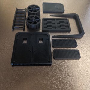 May include: A collection of black 3D-printed components, including a ladder, two fan-like structures, a door, and various rectangular and curved pieces. The parts are arranged on a light brown surface.