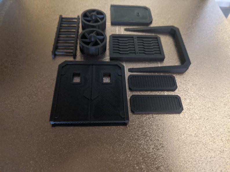 May include: A collection of black 3D-printed components, including a ladder, two fan-like structures, a door, and various rectangular and curved pieces. The parts are arranged on a light brown surface.