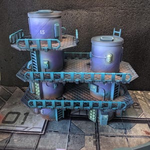 May include: A multi-tiered, miniature sci-fi structure with cylindrical tanks and platforms. The tanks are purple with the number "15" on one. The platforms and railings are blue-green. The model sits on a gray and red patterned surface.