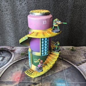 May include: A multi-level miniature building with a cylindrical design, painted in shades of purple, yellow, and green. The number "13" is visible on the side. Miniature figures are positioned on the structure, suggesting a tabletop game setting.