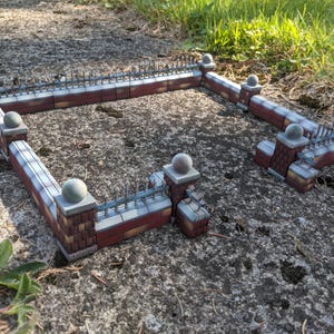 May include: A miniature brick wall fence section featuring red rectangular bricks, grey stone pillars topped with spherical finials, and grey metal spiked railings. The structure is arranged in a rectangular perimeter with an open gate section.
