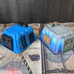 May include: Two miniature blue and gray sci-fi buildings. The blue building has a black door and ladder. The gray building has a gold-colored vent and green doors. Both have black vents on top. The buildings are on a gray and white surface.