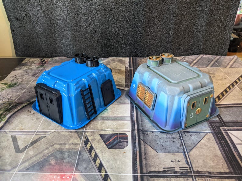 May include: Two miniature blue and gray sci-fi buildings. The blue building has a black door and ladder. The gray building has a gold-colored vent and green doors. Both have black vents on top. The buildings are on a gray and white surface.