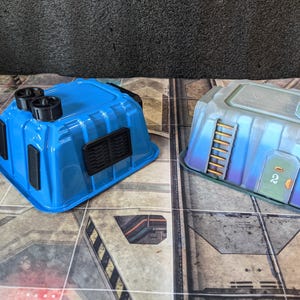 May include: Two miniature blue and gray terrain buildings for tabletop gaming. The blue building has black vents and circular details, while the gray building features a ladder, door, and propeller-like elements. Both are set on a patterned surface.