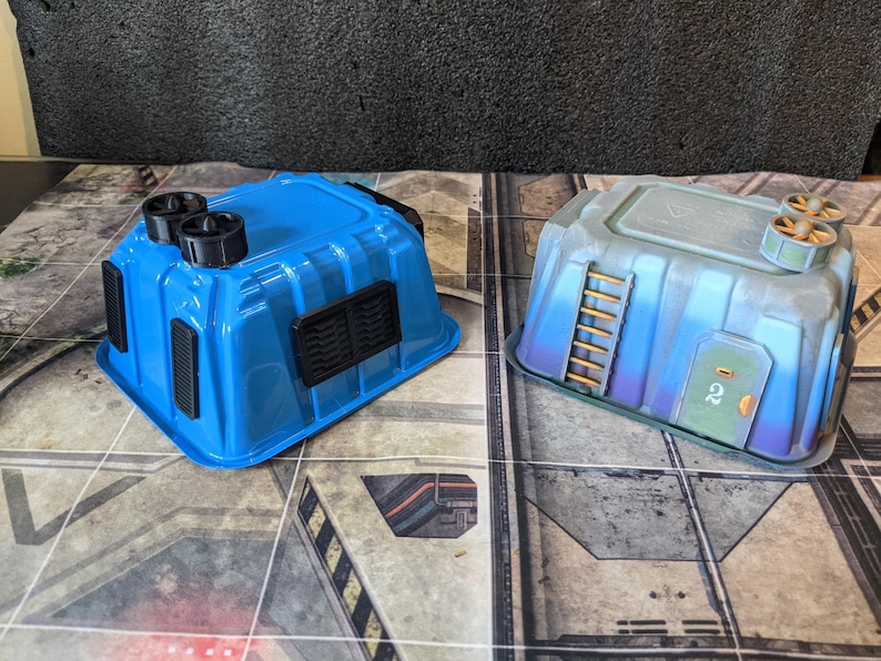 May include: Two miniature blue and gray terrain buildings for tabletop gaming. The blue building has black vents and circular details, while the gray building features a ladder, door, and propeller-like elements. Both are set on a patterned surface.