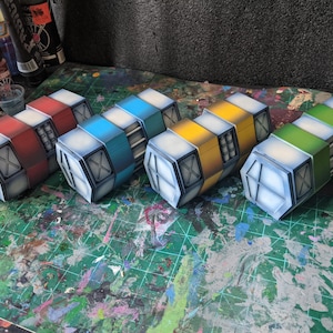 May include: Four rectangular sci-fi style containers arranged in a row, each featuring a distinct color scheme of red, blue, yellow, and green. The containers have white end caps with black cross-hatch detailing and a central section with vertical bars.