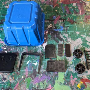 May include: A blue plastic container and several black plastic model parts are arranged on a green cutting mat. The parts include panels, a ladder, and fan-like components, likely for a model or miniature project.