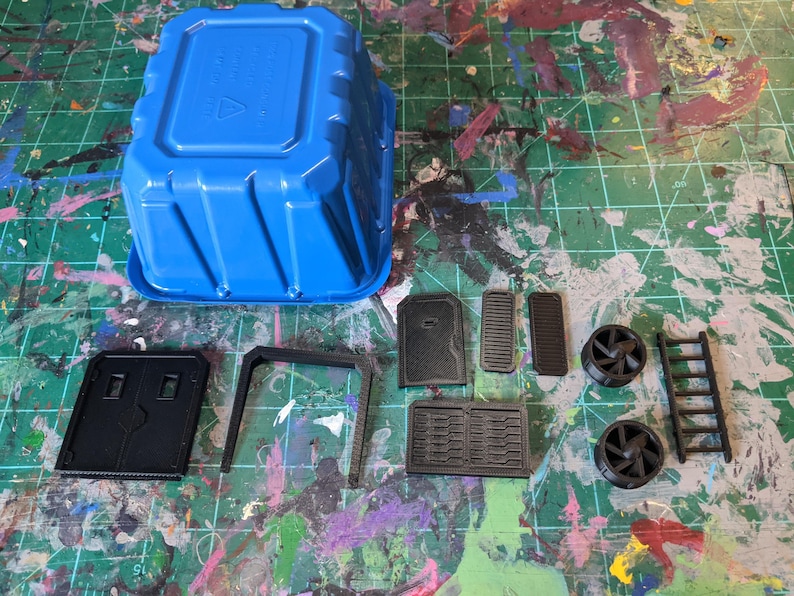 May include: A blue plastic container and several black plastic model parts are arranged on a green cutting mat. The parts include panels, a ladder, and fan-like components, likely for a model or miniature project.