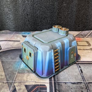 May include: A miniature, futuristic-style building with a gray and blue color scheme. The structure features two small ventilation units on top, two green doors with the number "2", and a small orange ladder. The building is set on a patterned surface.
